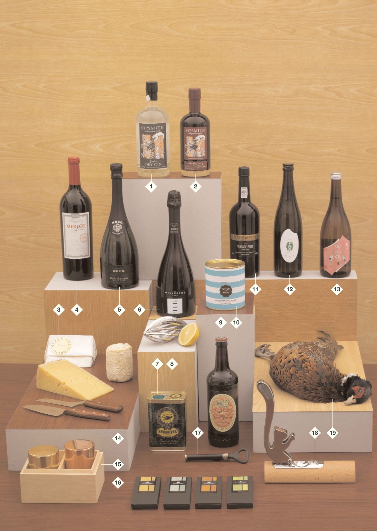 Styled display of food and drink gift items including wine bottles, cheese, spirits, tea caddies, and kitchen tools on wooden platforms.