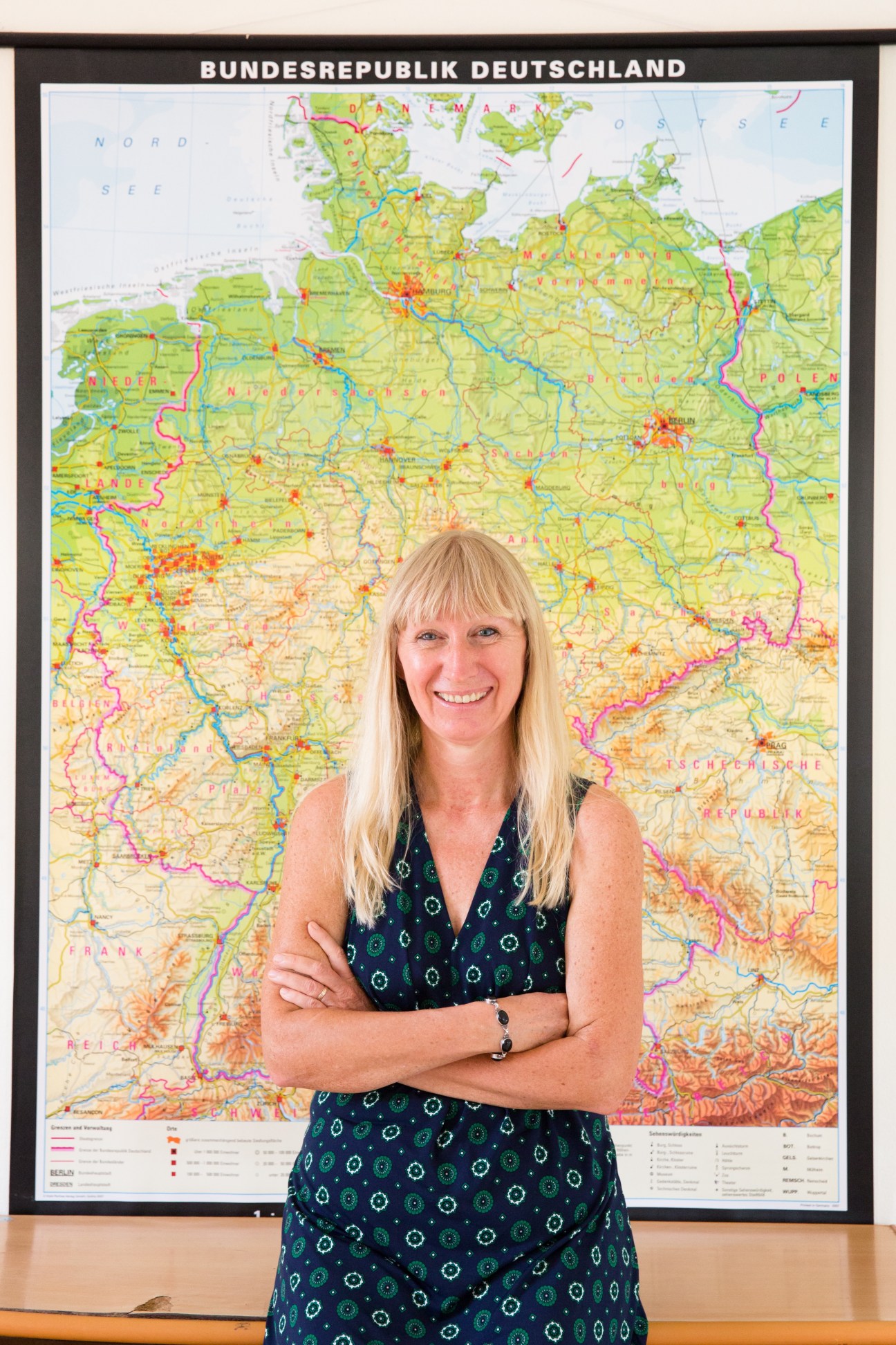 Teacher Rita Schneider smiles at the camera, standing with crossed arms in front of a large colorful map of Germany labeled Bundesrepublik Deutschland.