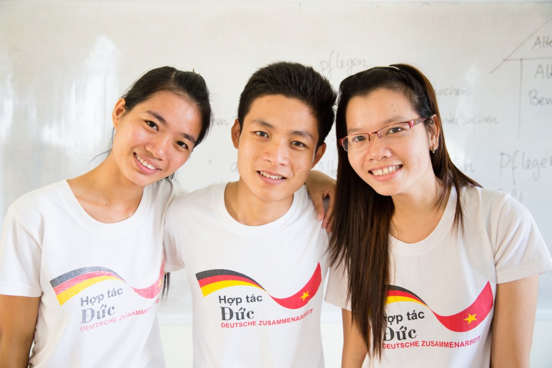Three smiling Vietnamese nursing students wearing white t-shirts with German and Vietnamese flags displaying Vietnamese-German cooperation.