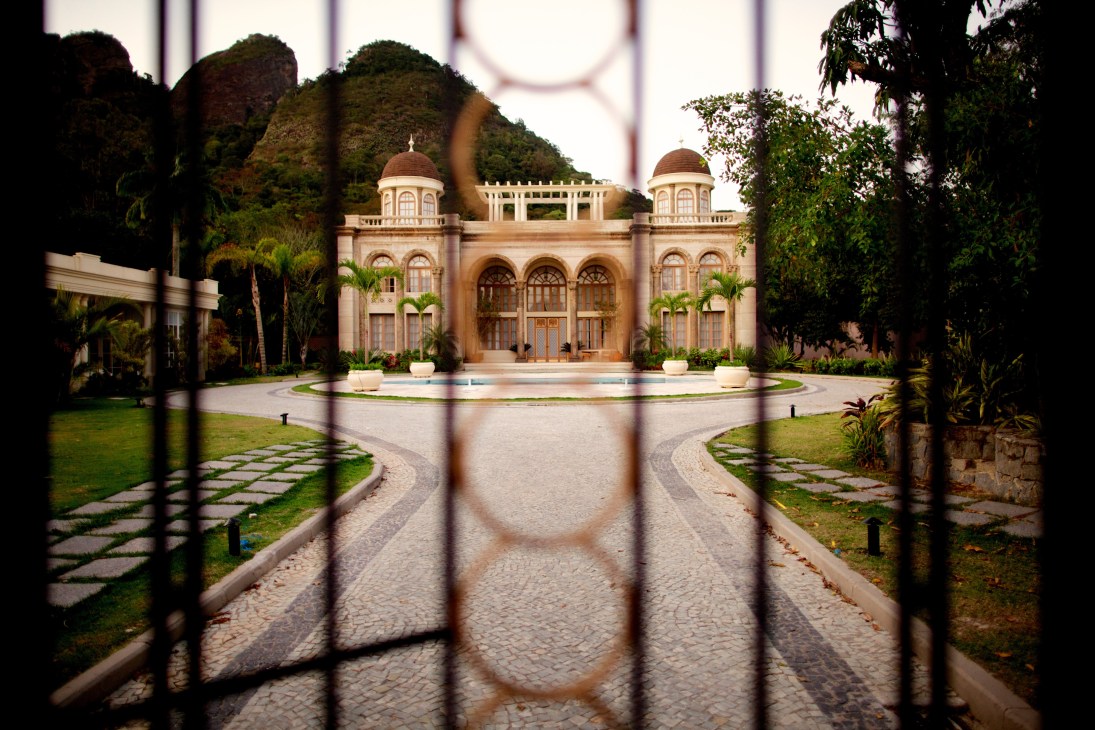 Ornate mansion set with domed towers and manicured grounds viewed through a decorative metal fence.