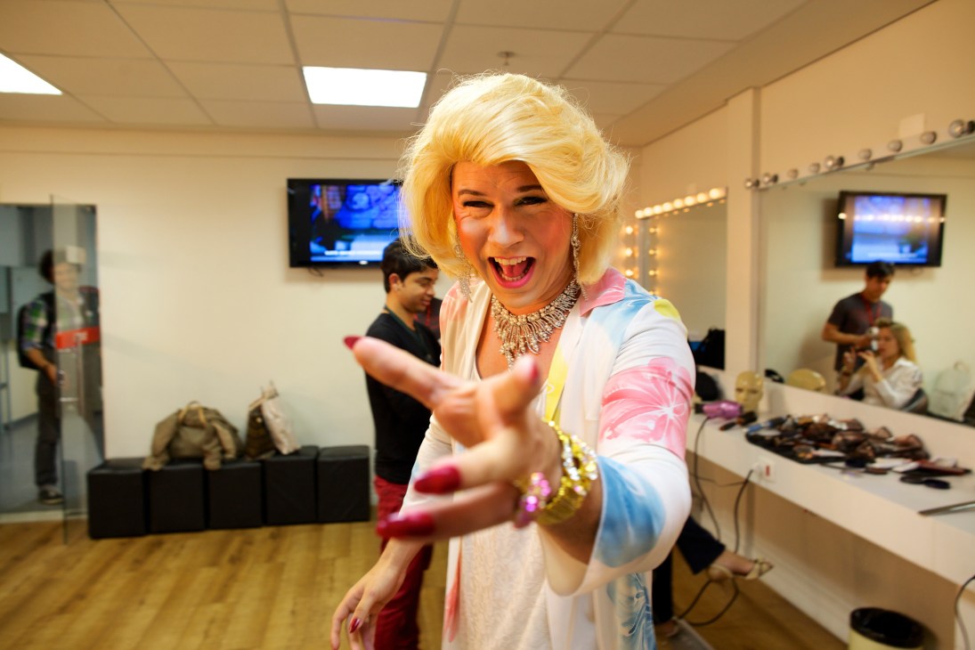 Drag performer in blonde wig and colorful outfit poses in dressing room with mirrors and lights.
