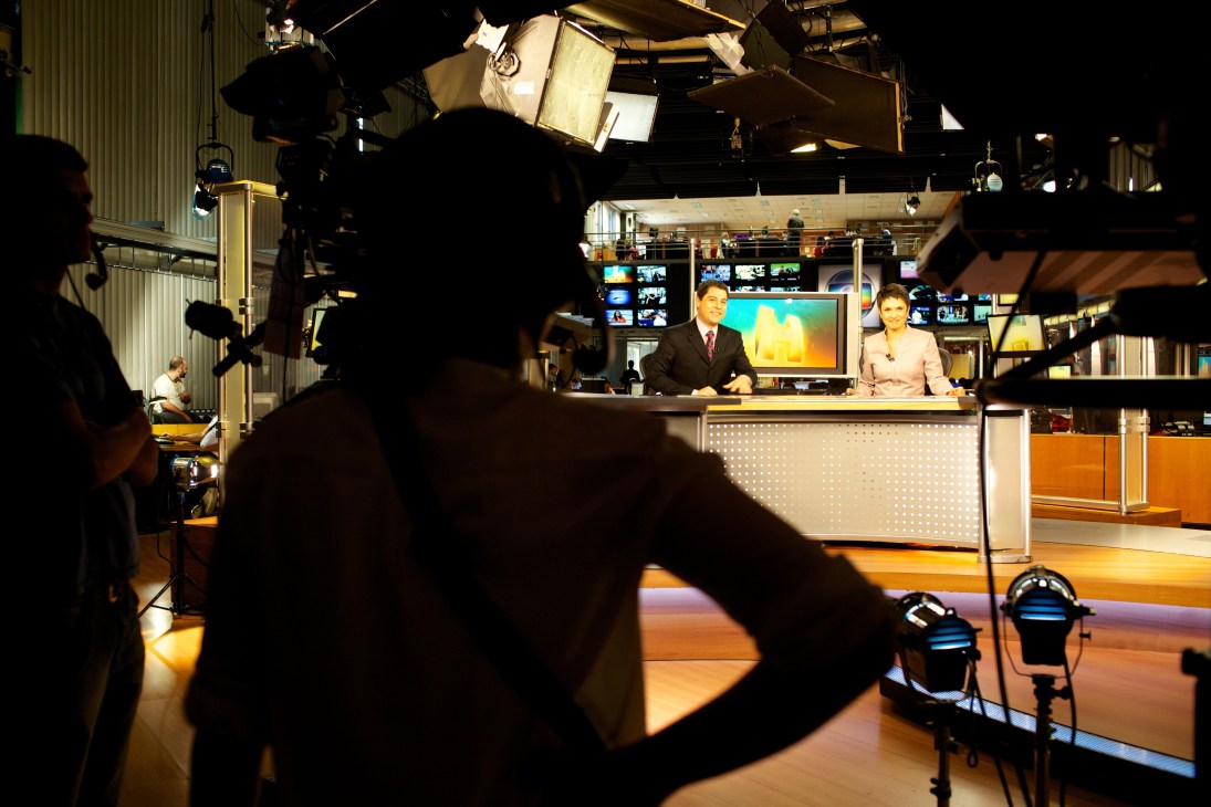 Two news anchors sit at a desk in a broadcast studio with professional lighting and monitoring equipment visible in the background.