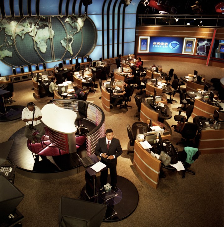 Overhead view of Record's São Paulo newsroom with journalists working at curved wooden desks under world map displays.