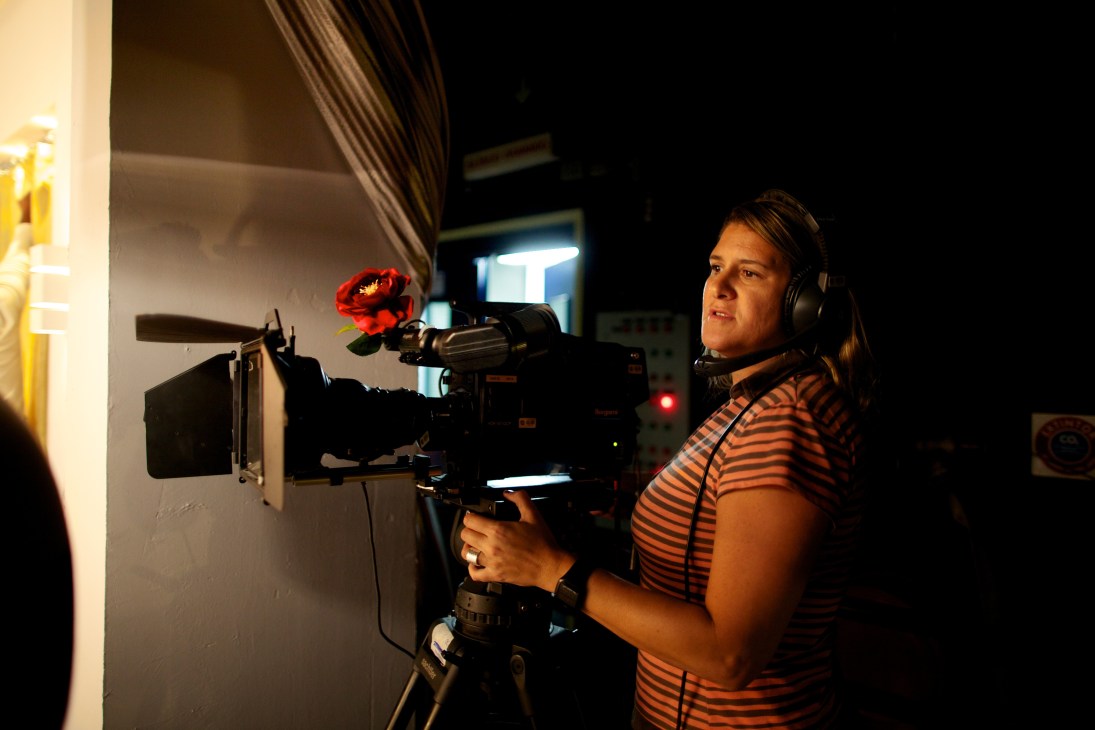 Female camera operator in striped shirt operating professional video camera with red microphone attachment.