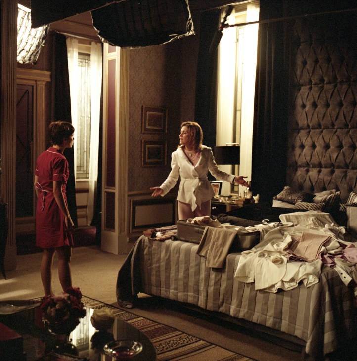 Two women in a bedroom set on a television production stage, one in white blazer and one in red dress.
