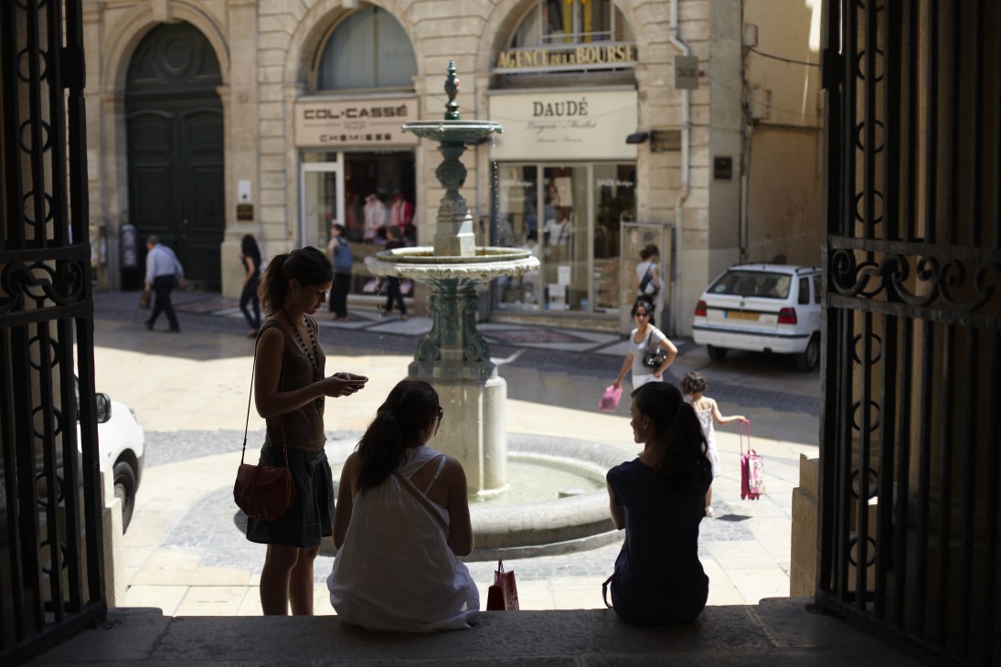 Three women sit in a shaded doorway overlooking a sunny plaza with a central fountain and historic sandstone buildings in Montpellier's old town.