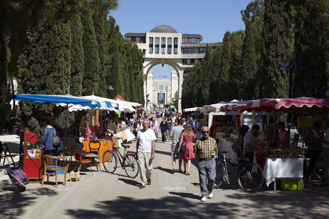 Market stalls with colorful umbrellas line a tree-lined pedestrian avenue in Montpellier's Antigone district.