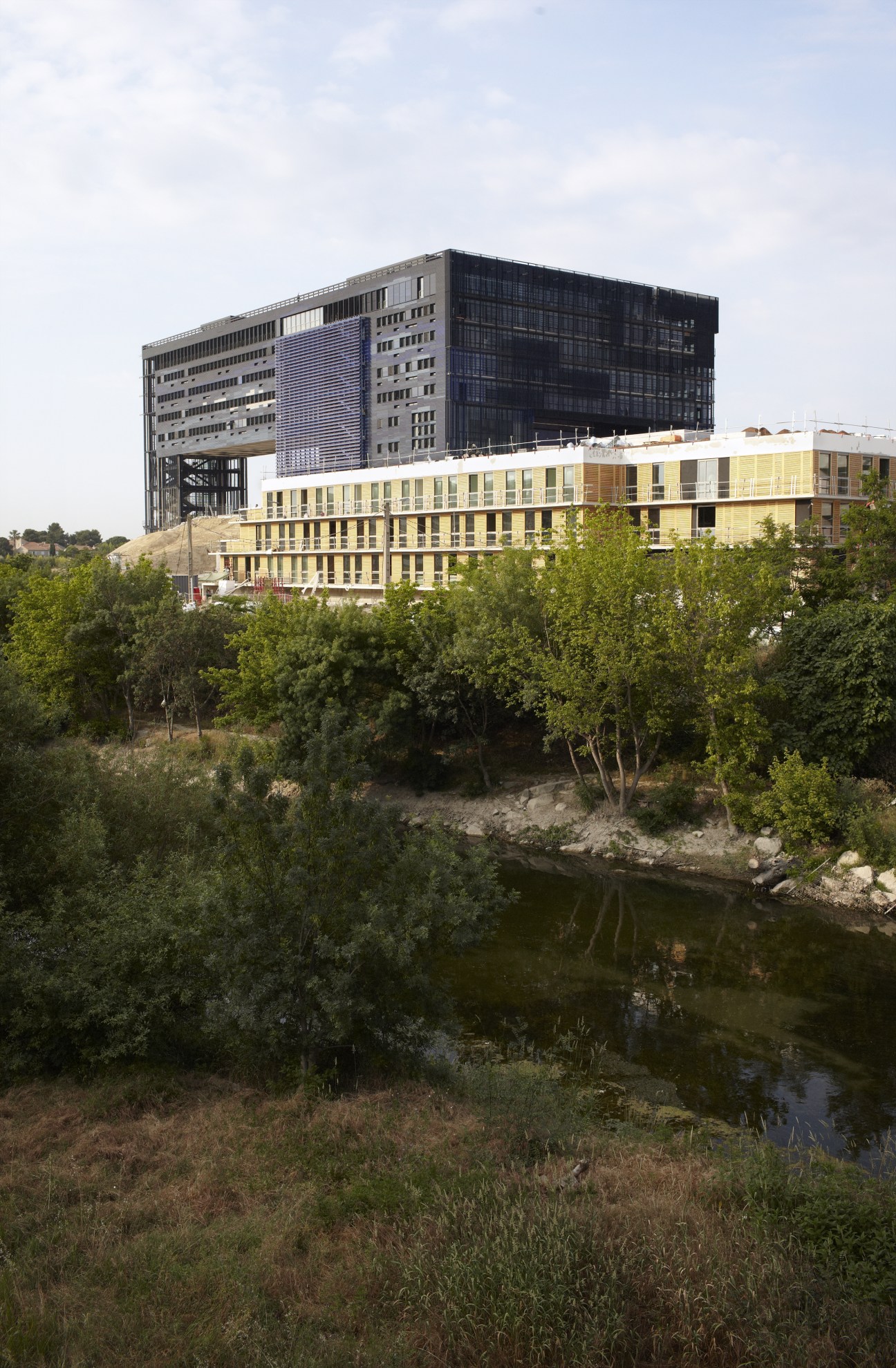 Modern yellow building with dark upper structure beside a river surrounded by green vegetation.
