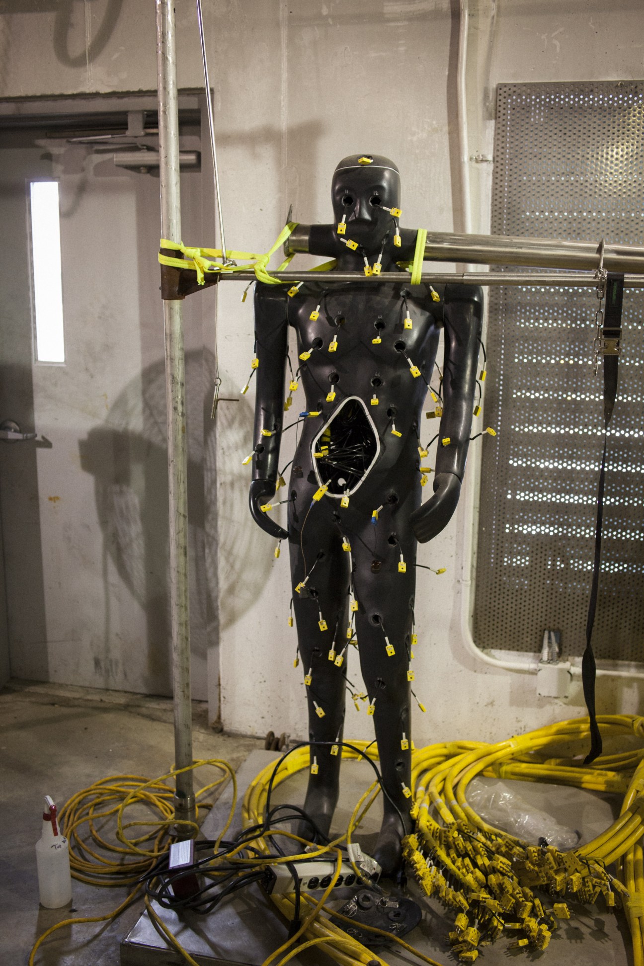 Black mannequin covered in yellow heat sensor patches and wires in a laboratory testing facility.