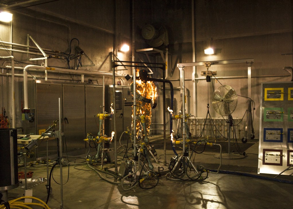 Fire testing equipment with flames engulfing a mannequin wearing military combat uniform in a laboratory setting.