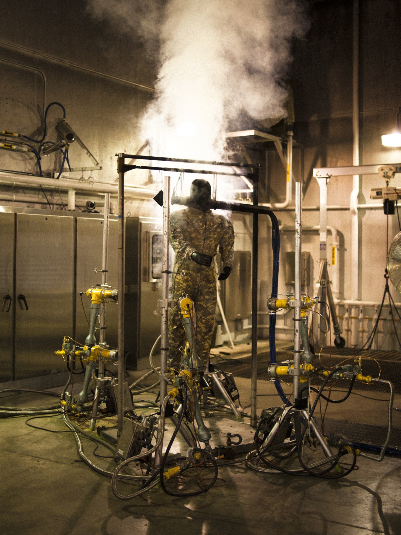 Military test dummy in camouflage uniform surrounded by heat testing equipment with smoke rising above.