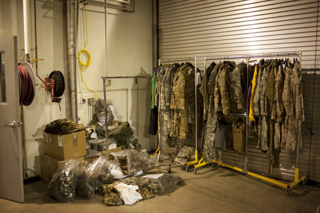 Military uniforms and camouflage fatigues hang on racks in a testing facility, with plastic bags of fabric samples on the floor.