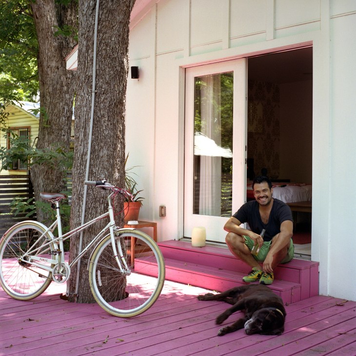 Alan Gonzalez sits on a pink porch with his black labrador dog Taco, a fixed-gear bike leaning against a tree nearby.