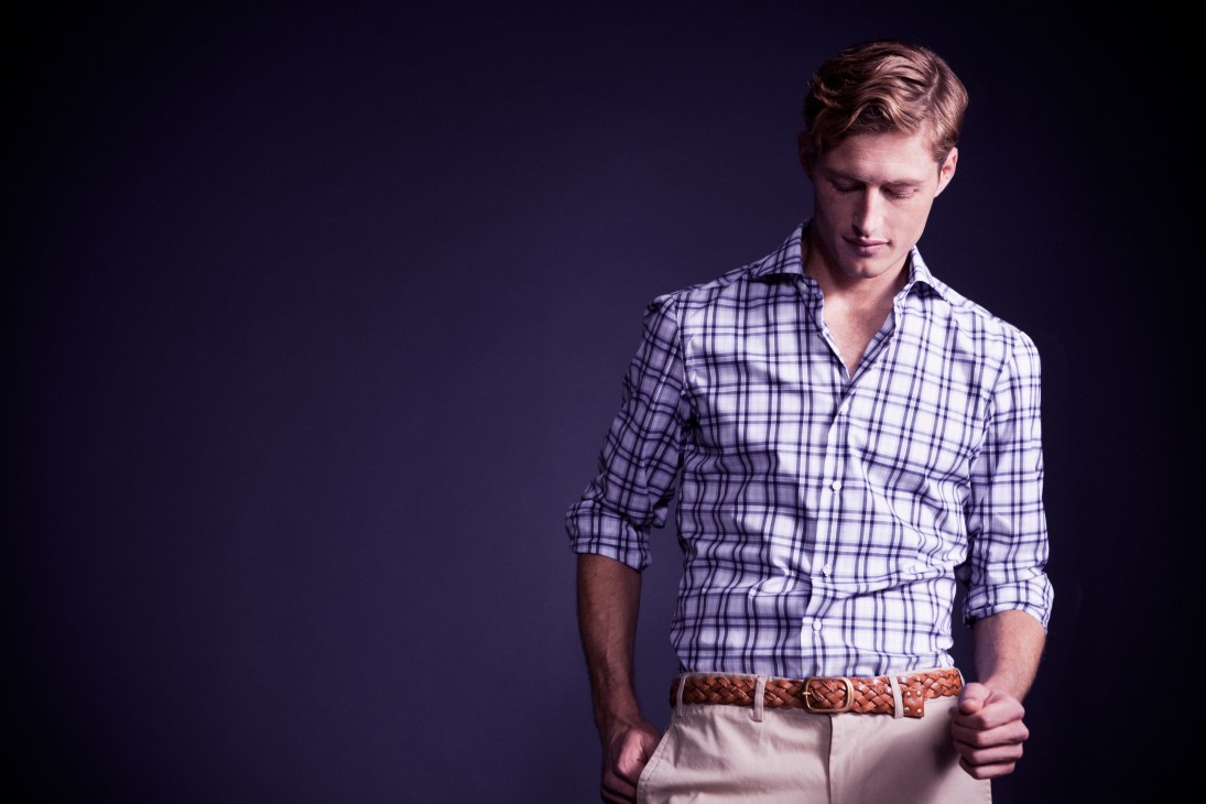 Man wearing purple check shirt and beige trousers with braided belt against dark background.