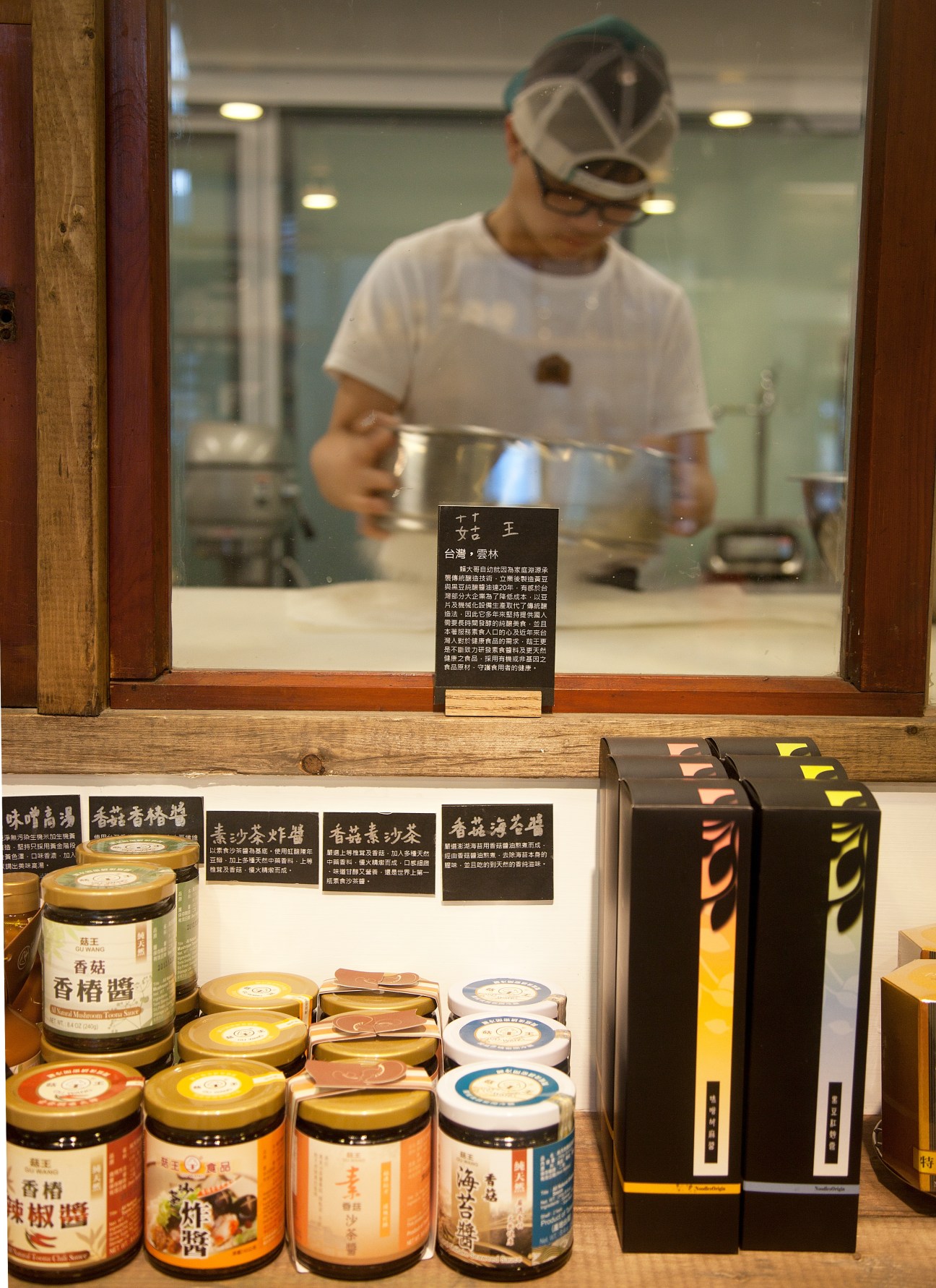 A person in a cap prepares food at Good Cho's café counter in Taipei, viewed through a window display of packaged goods and jars.