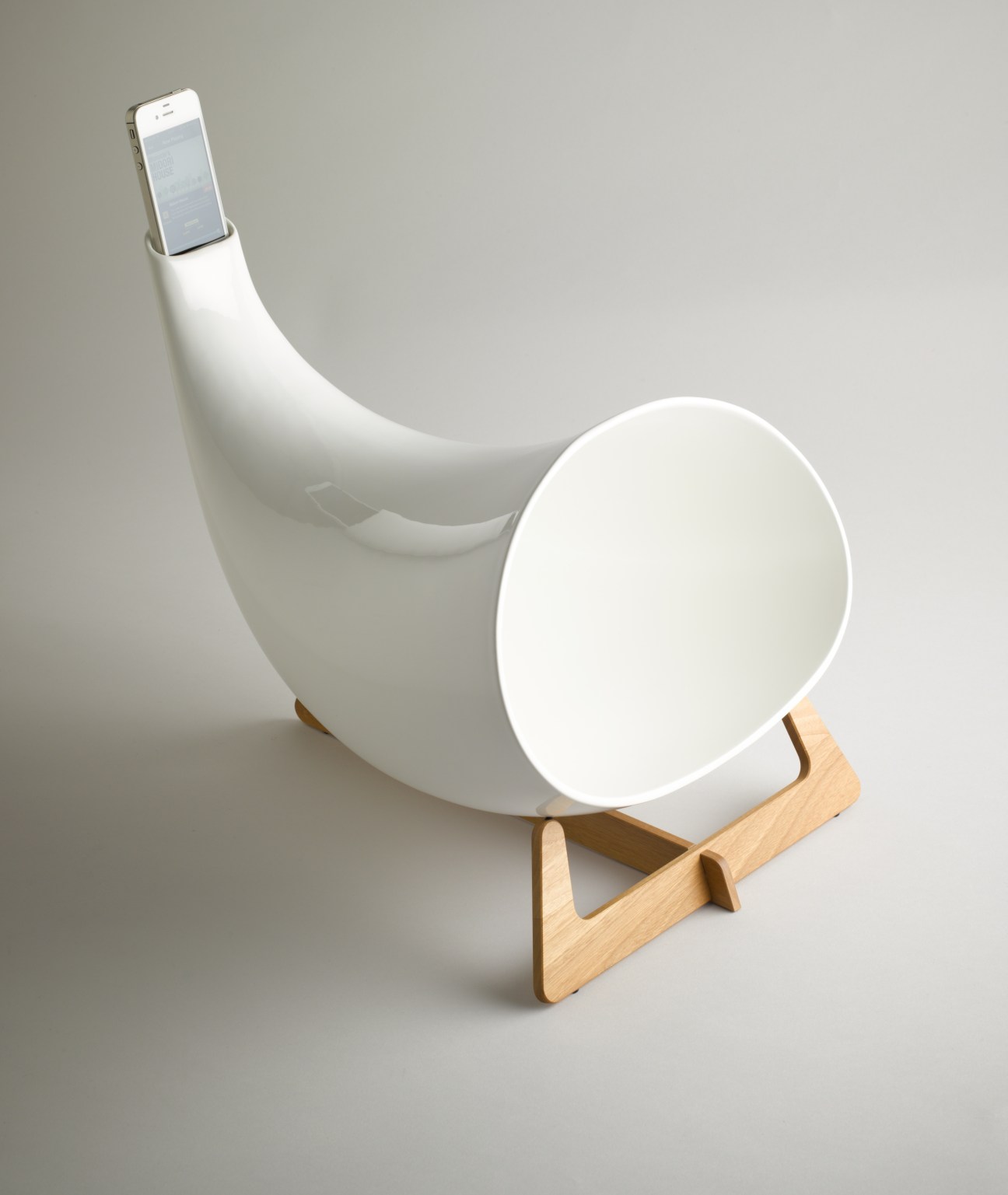 White ceramic horn-shaped speaker with iPhone inserted at the top and oak tripod stand supporting the base.