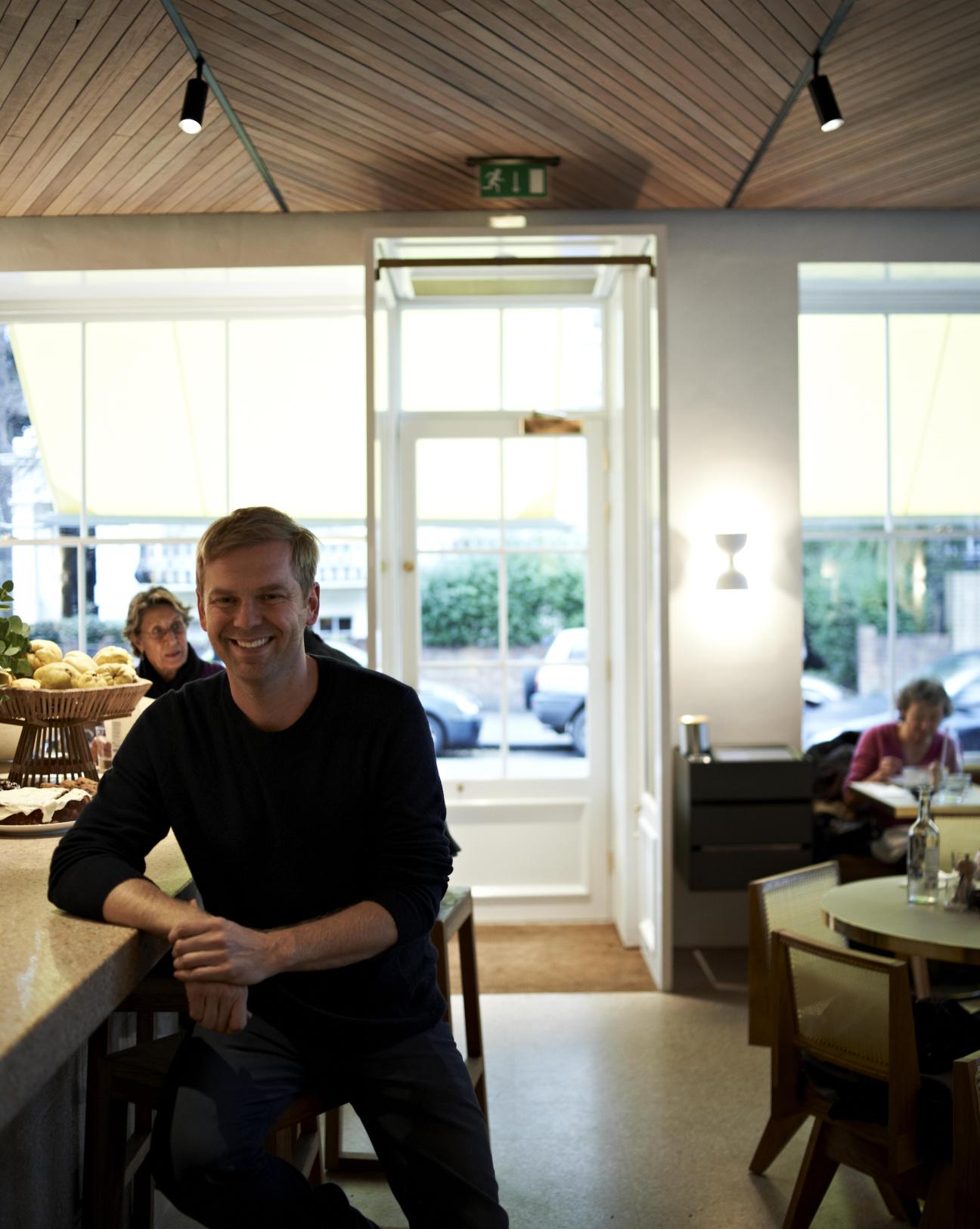 Bill Granger smiles at the counter of his Granger & Co restaurant in London, with large windows and customers visible in the bright, modern interior.