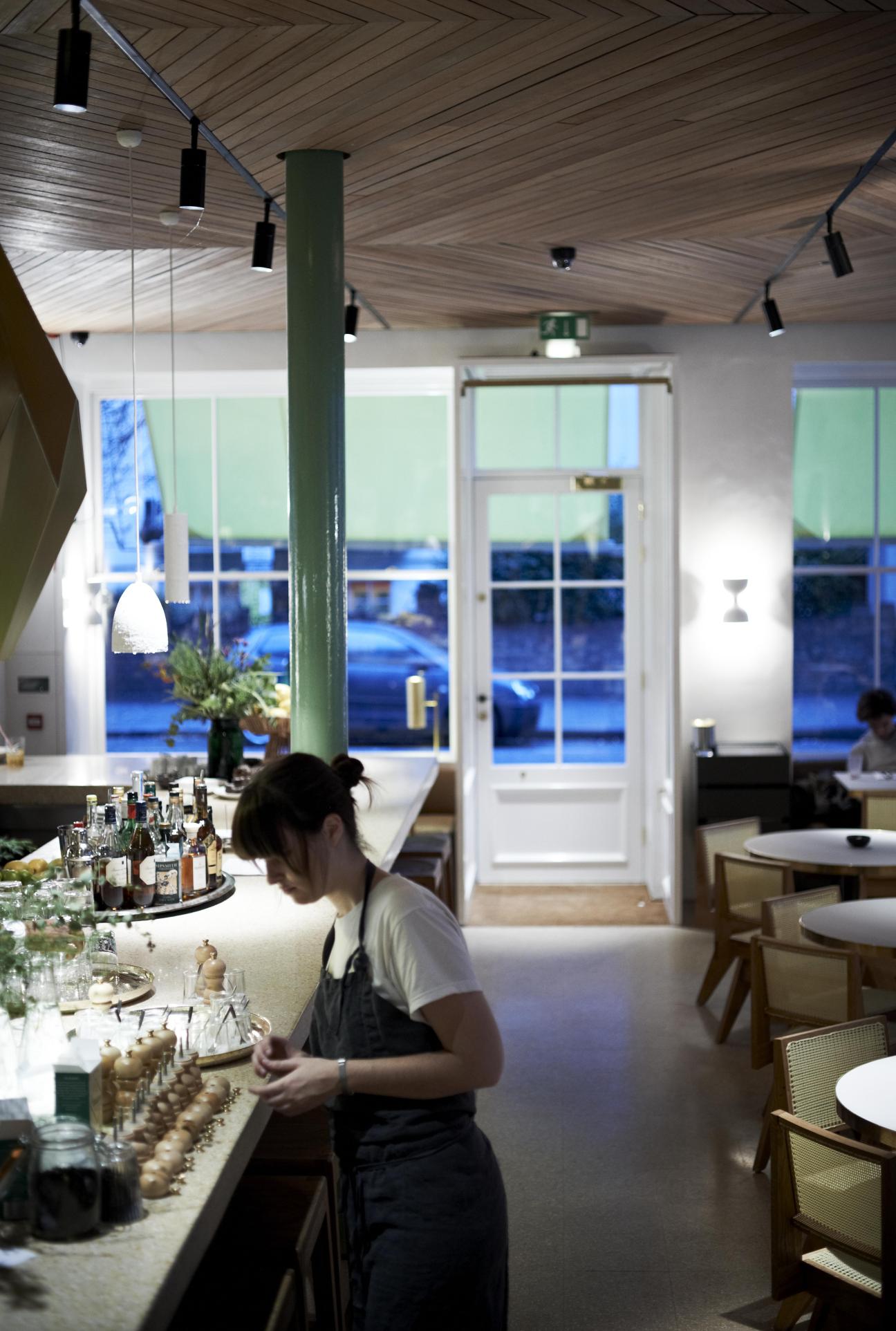 Staff member at Granger & Co restaurant bar with bottle display and large windows overlooking waterfront.