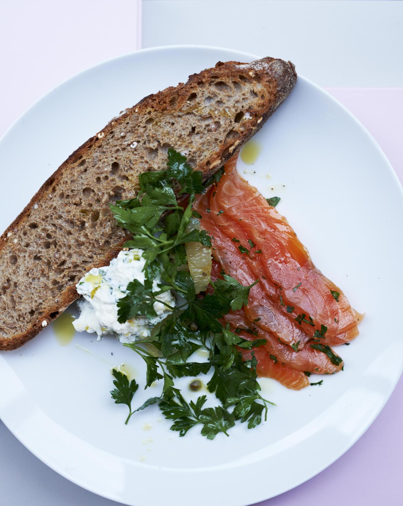 Slice of toasted whole grain bread topped with smoked salmon, cream cheese and fresh parsley on a white plate.