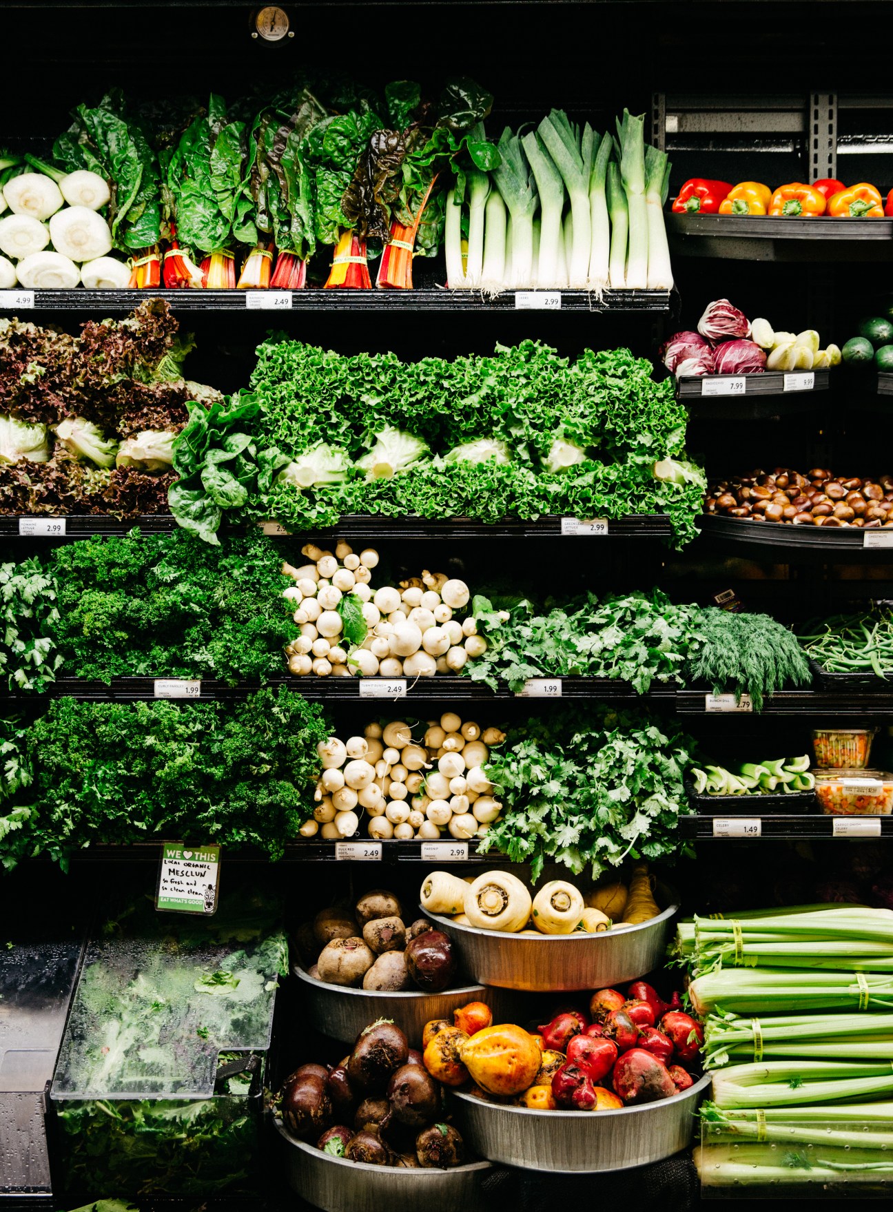 Tiered black shelves displaying fresh organic vegetables including lettuce, peppers, onions, and leafy greens with price tags.