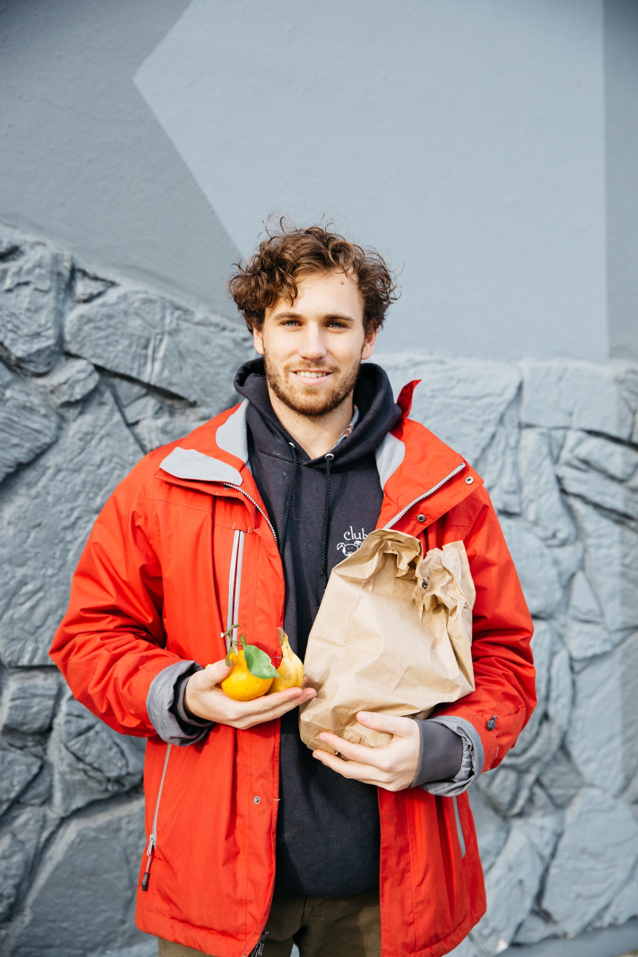 Man in red jacket holding fresh lemons and paper bag against textured wall