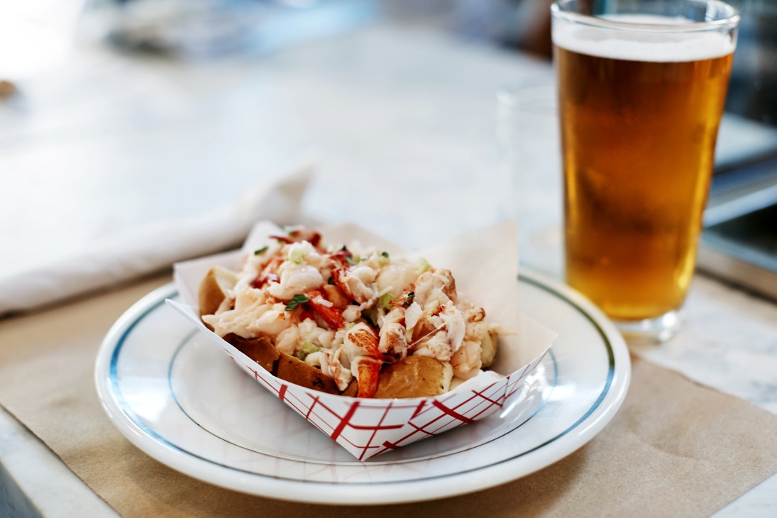 A lobster roll served on a grilled bun with tarragon and lemon, accompanied by a glass of beer.