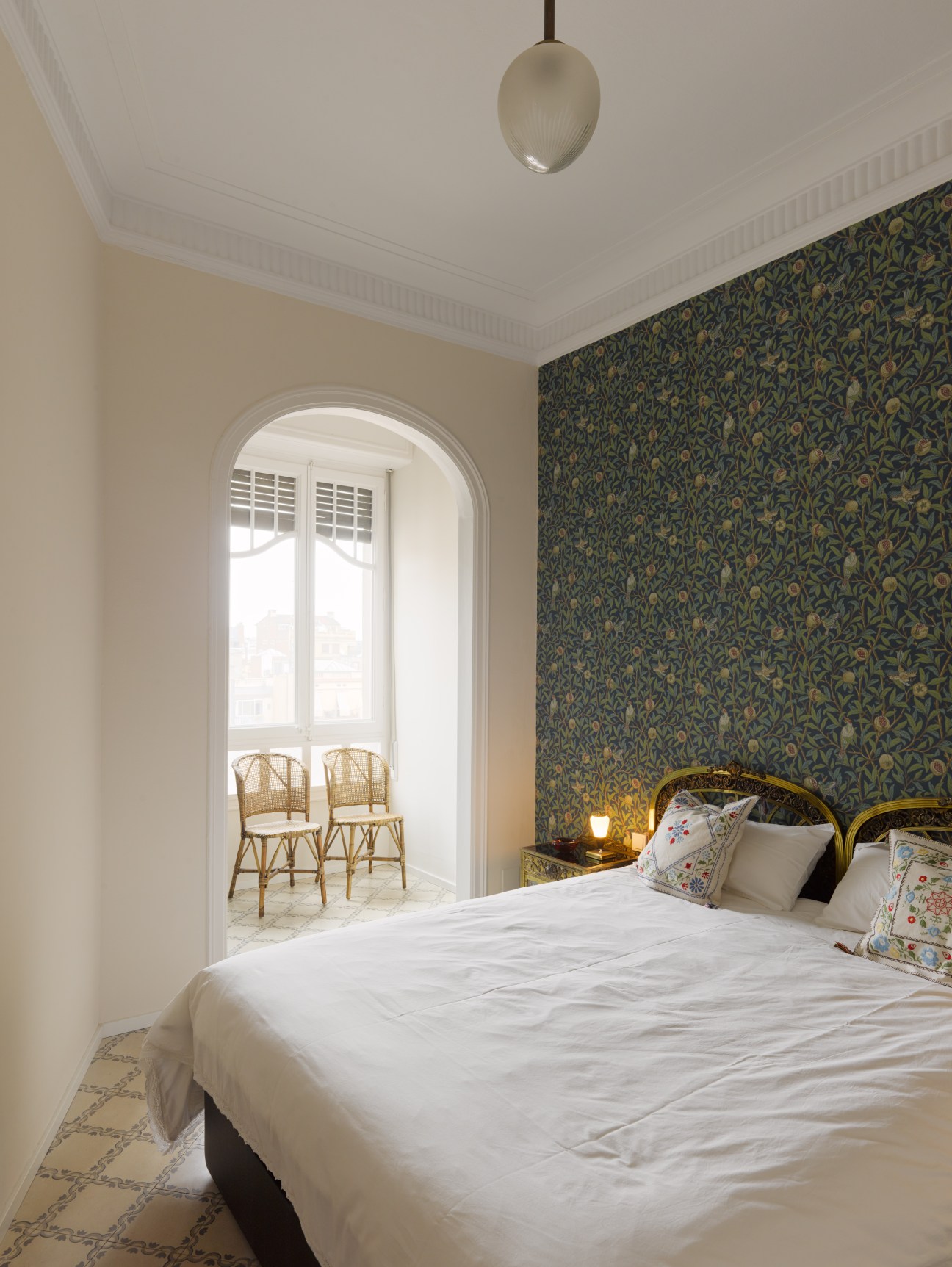 Bedroom with patterned emerald wallpaper, gold-framed bed, white linens, and arched doorway to balcony with rattan chairs.