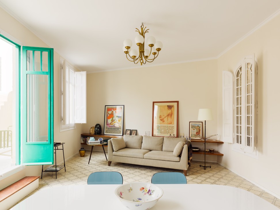 Minimalist lounge with cream sofa, turquoise chairs, brass chandelier, and framed artwork on pale yellow walls.