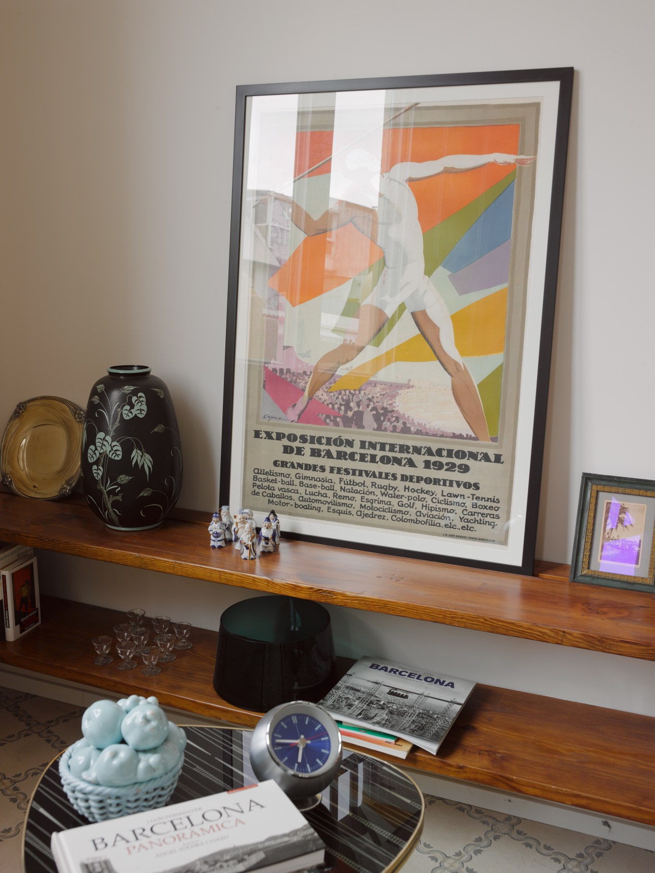 Wooden desk displaying a framed 1929 Barcelona sports exposition poster, decorative vases, clock, and design books.