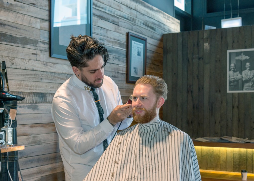 A barber in a white shirt and tie trims a customer's beard at Groomed salon in Braddon.