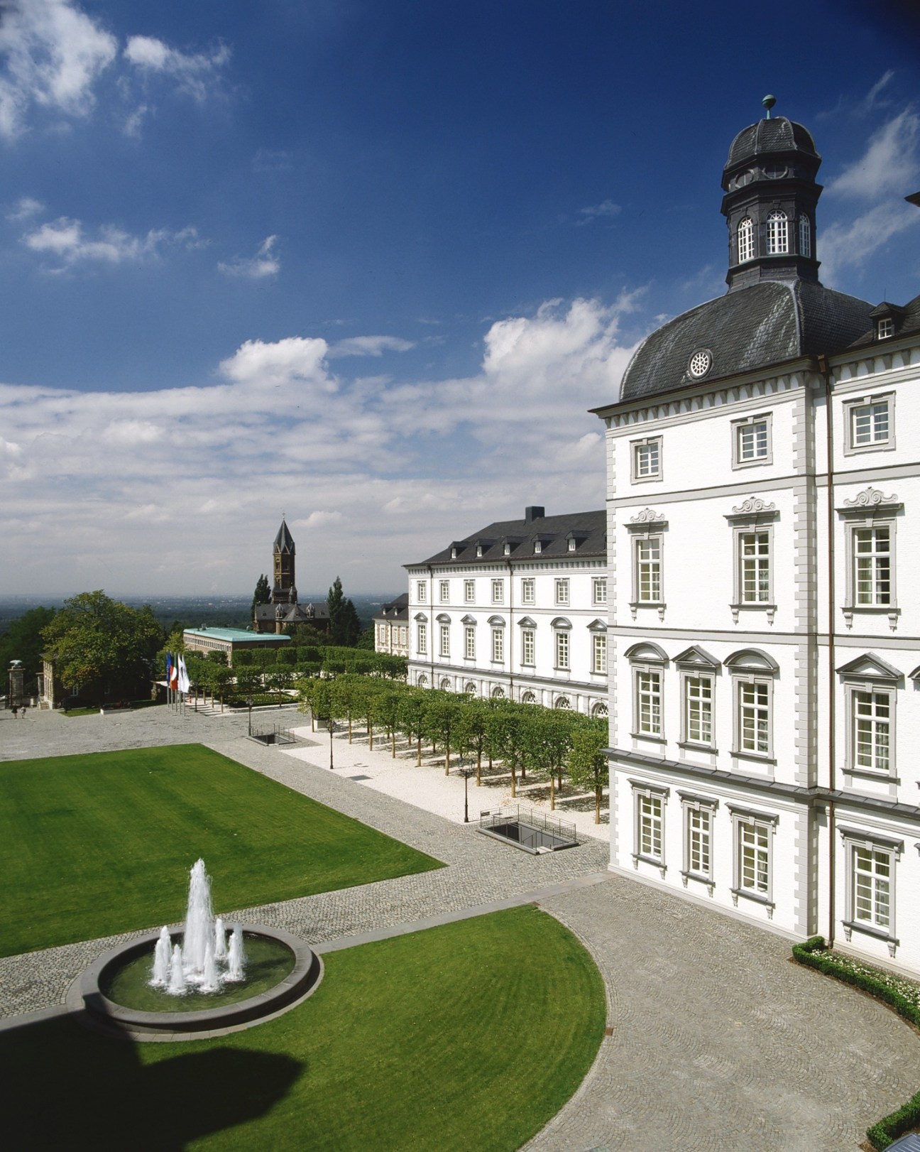 Exterior view of Grandhotel Schloss Bensberg, a white neoclassical building with a domed clock tower overlooking manicured gardens and distant landscape.