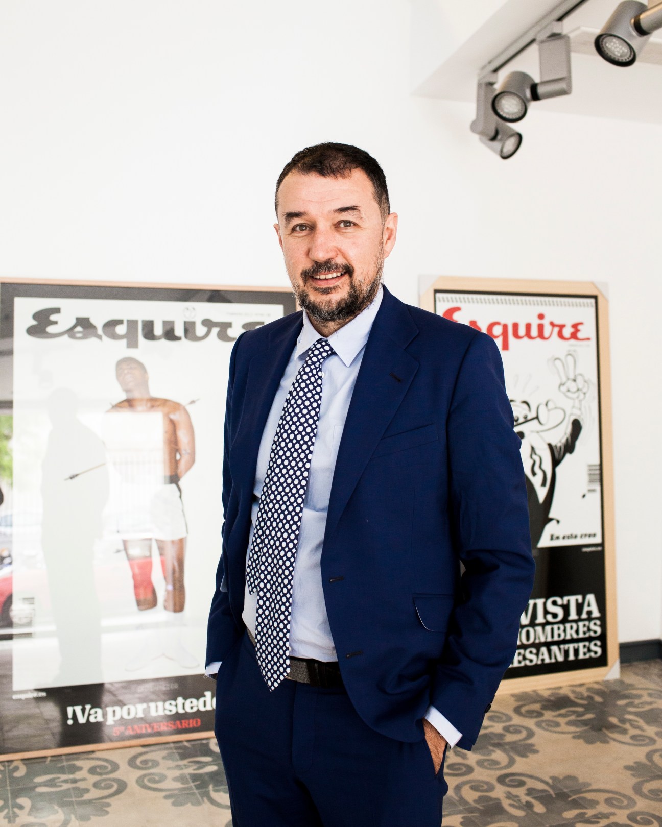 Man in navy blazer and patterned tie stands in office with Esquire magazine covers on wall behind him.