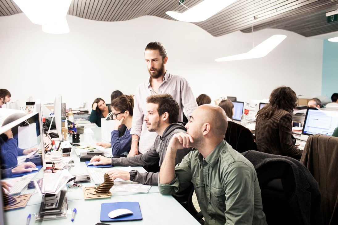 Team members gathered around a white desk in a bright, modern office space with corrugated steel ceiling.