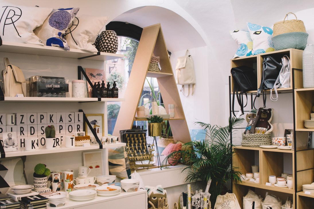 Interior of Gud design shop displaying handmade Slovenian products including ceramics, leather bags, wooden jewelry, and woven baskets on white shelves.