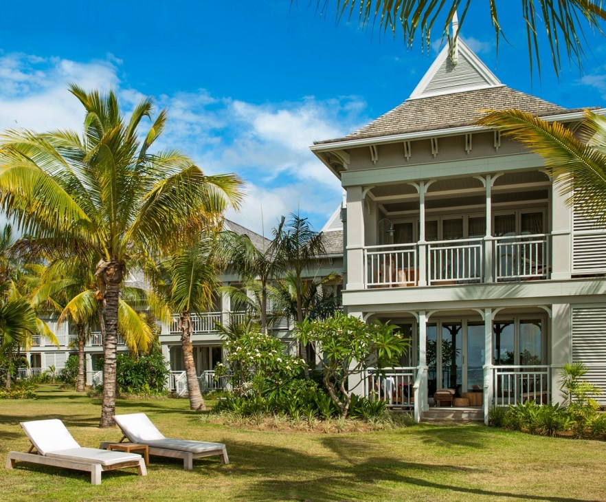 White colonial-style villa with palm trees and lounge chairs on manicured lawn under blue sky.