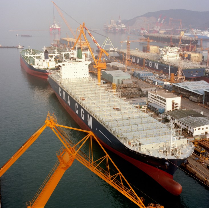Large container ship under construction at DSME shipyard with orange cranes and industrial facilities visible.