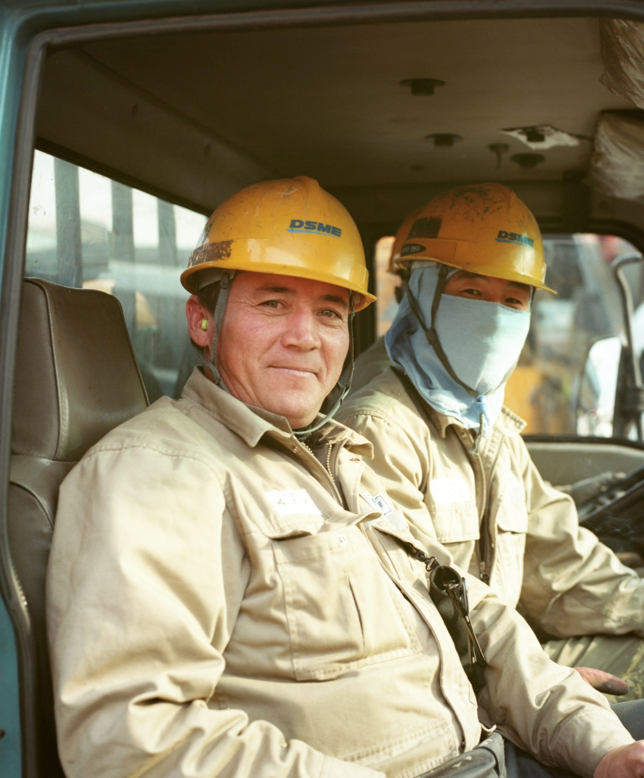 Two DSME workers wearing yellow hard hats and protective gear sit in a vehicle at the shipyard.