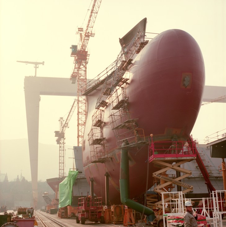 Large red spherical section of a container ship under construction at DSME shipyard with industrial cranes and scaffolding.