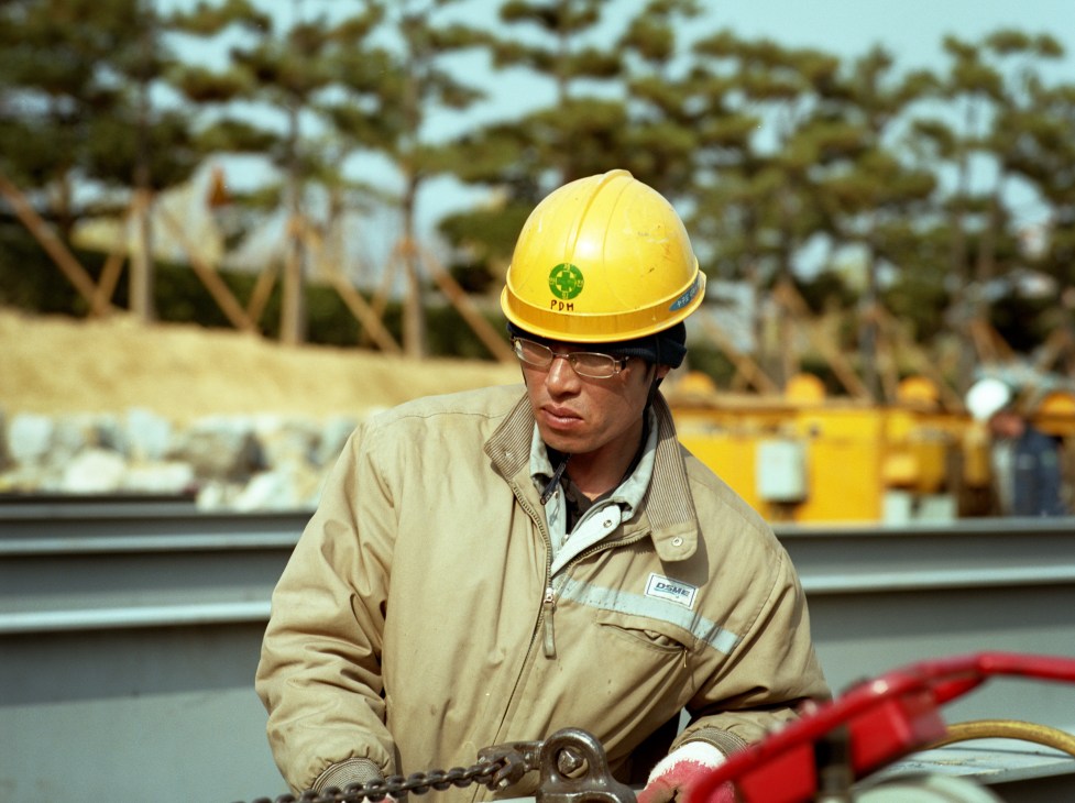 DSME worker wearing yellow hard hat and beige jacket on a ship deck.