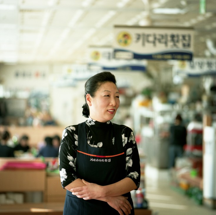 Woman in black floral apron smiling at Jagalchi fish market in Busan, Korea.