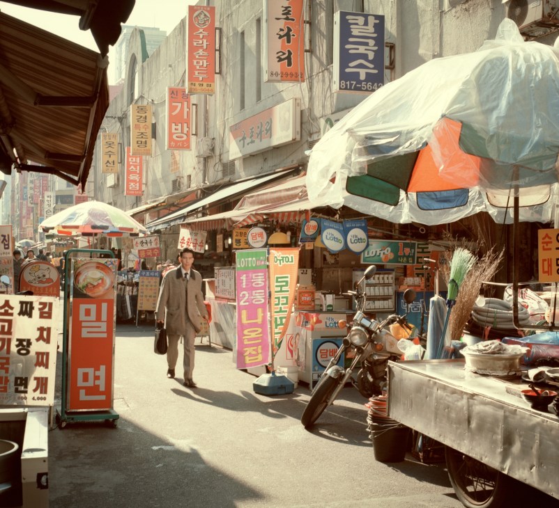 A busy shopping street in Busan with Korean signage, market stalls with white awnings, and a man in a brown coat walking through the crowded marketplace.