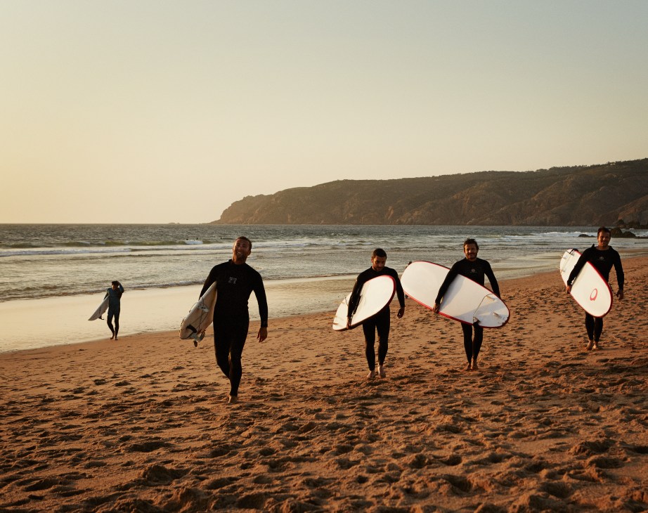 Surfers in wetsuits carrying white and red surfboards walk out of the sea onto Guincho Beach's sandy shore.