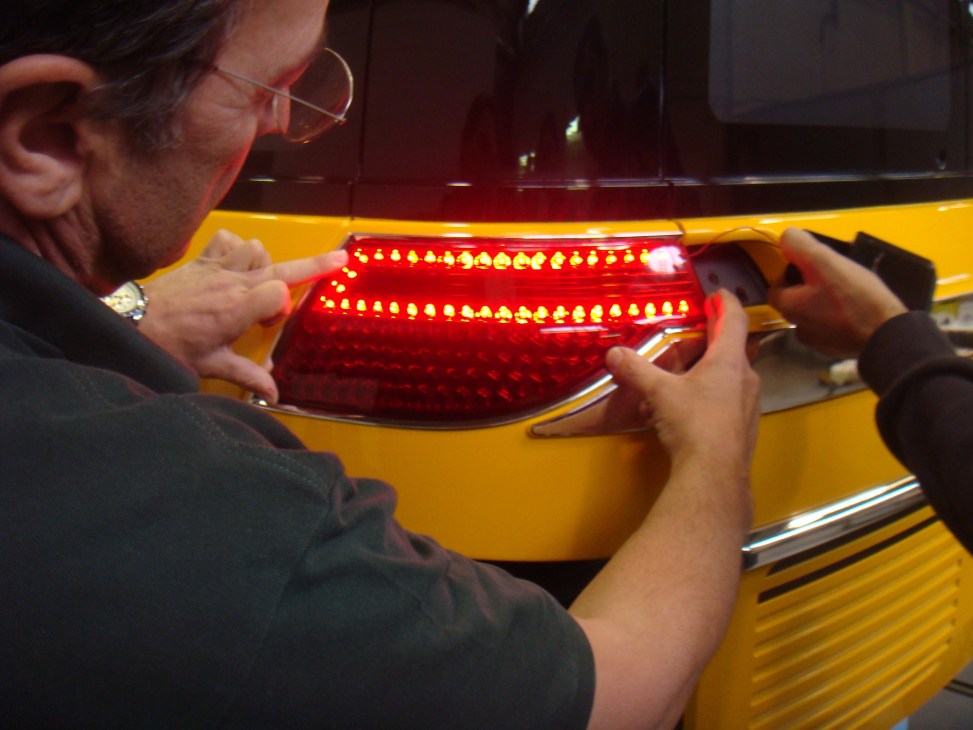 Man inspecting red LED taillight on yellow Karsan V-1 taxi prototype.