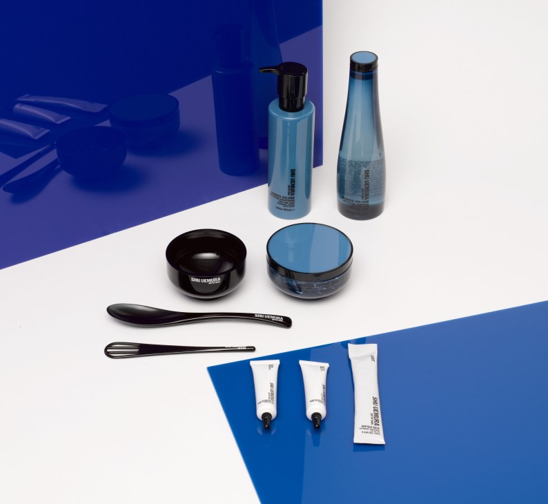 Shu Uemura hair care products arranged on a blue and white surface, including bottles, jars, applicators, and sample tubes.