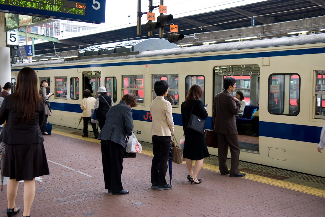 Passengers board a blue and cream JR train at a Japanese railway station platform.