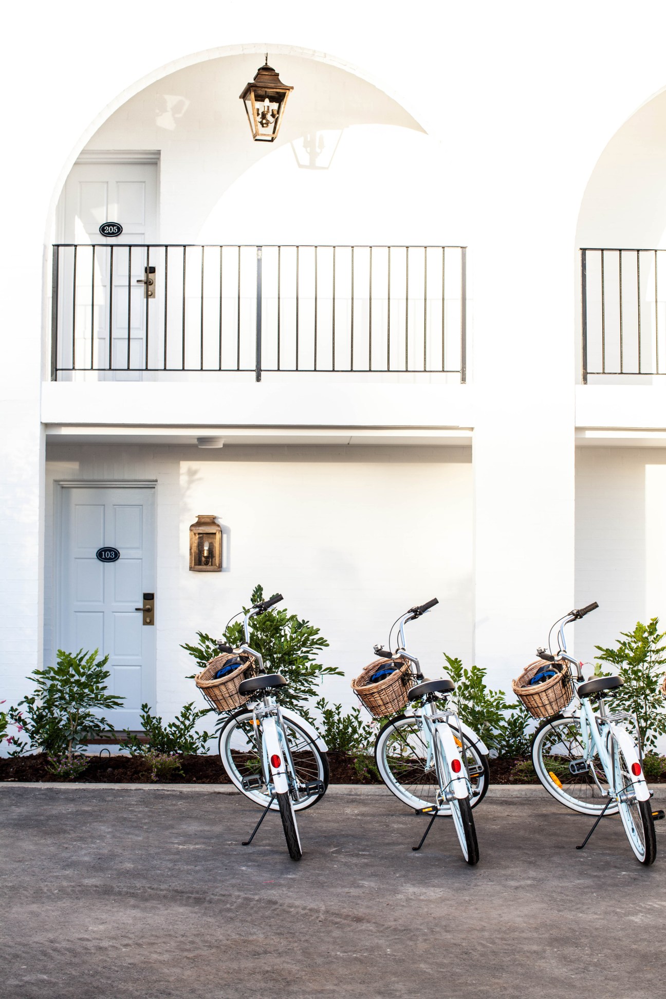 Three white bicycles with wicker baskets parked in front of a white building with arched architecture and room numbers.