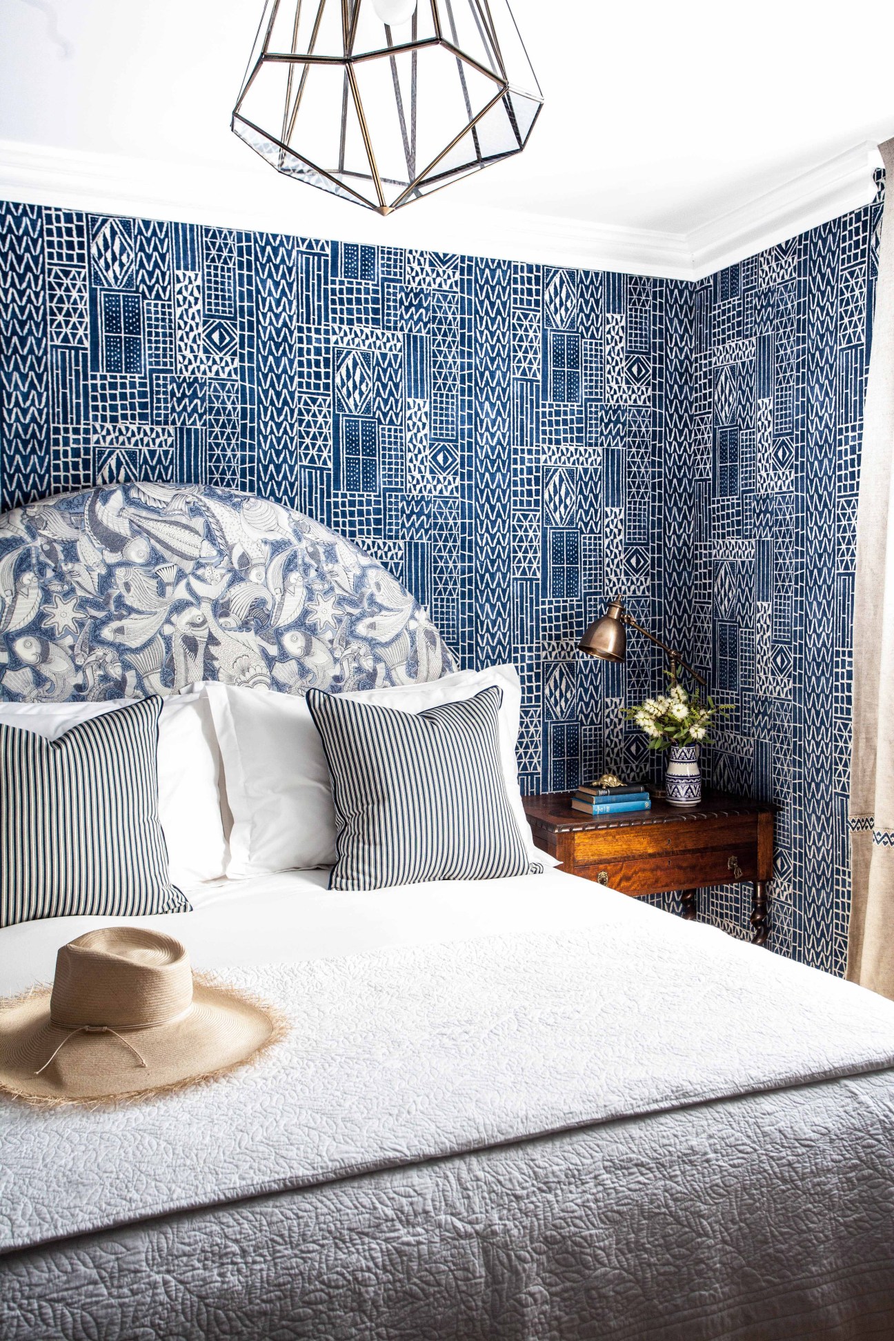 A boutique hotel bedroom with blue patterned wallpaper, white bedding, striped pillows, geometric pendant light and wooden nightstand.
