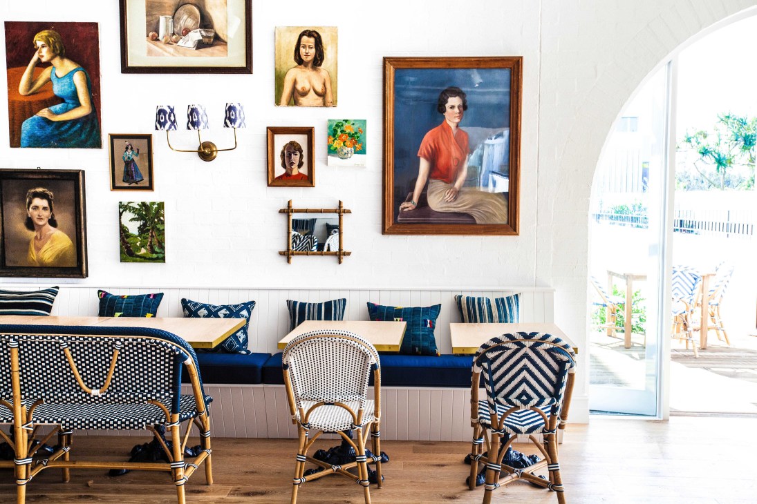 Interior seating area with patterned rattan chairs, blue cushioned bench, and gallery wall of framed artwork and portraits.