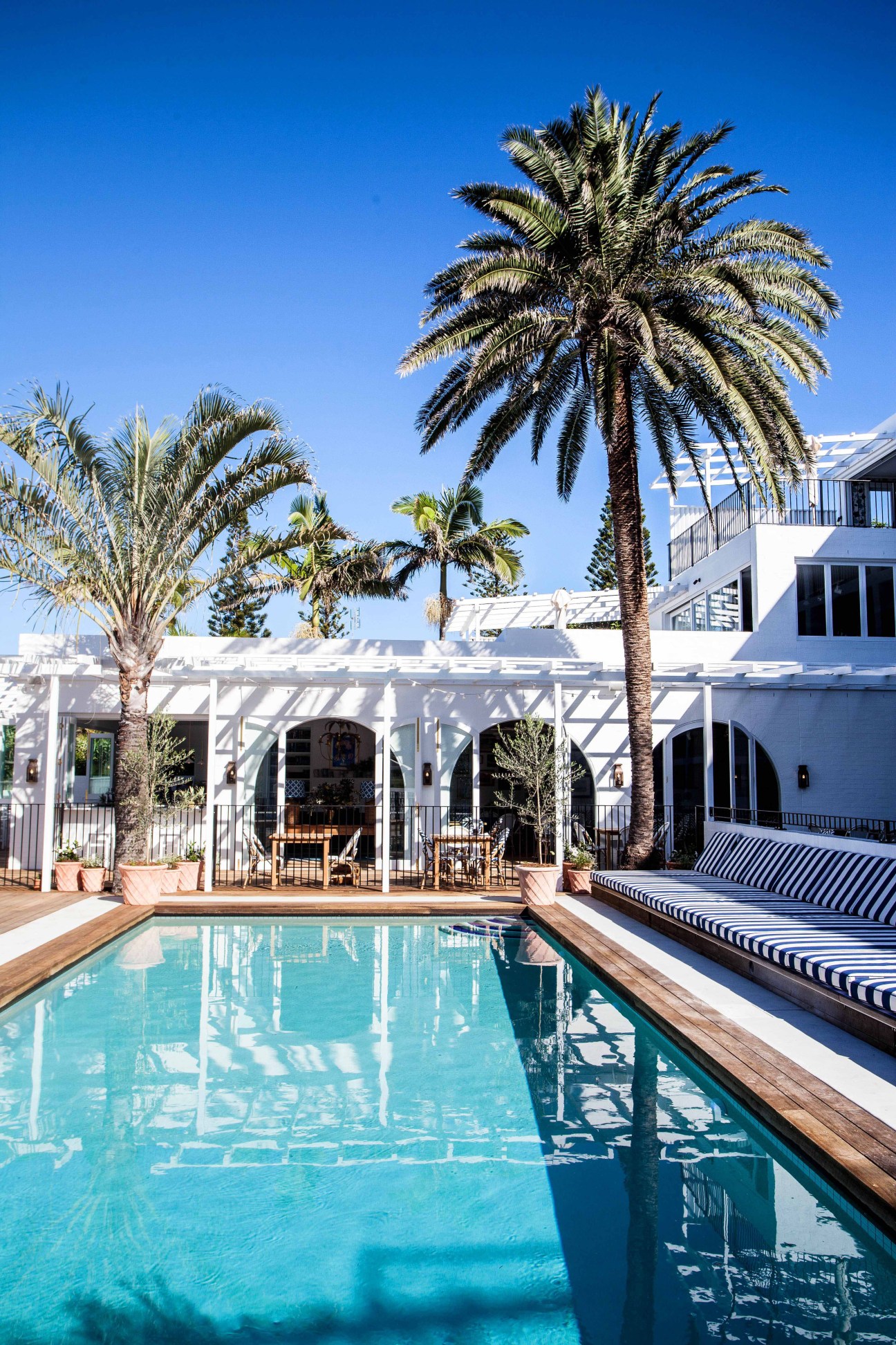 Halcyon House poolside with turquoise water, palm trees, white pergola and striped loungers under blue sky.