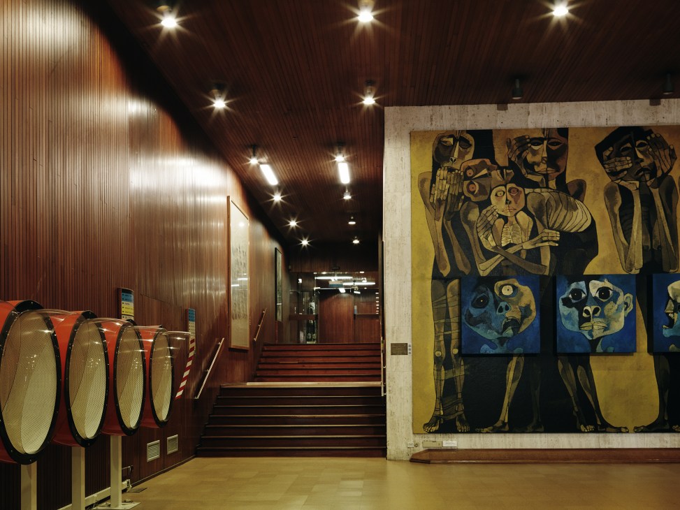 Modernist corridor with wood paneling, recessed lighting, sculptural red installation, and figurative artwork on concrete wall.